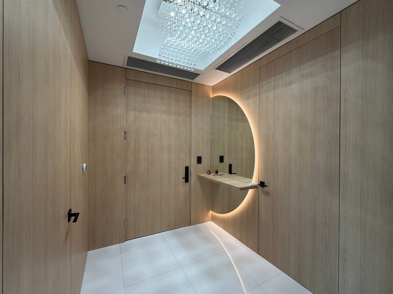 Entry vestibule clad in light wood-grain panels with flush doors, a backlit round mirror, a floating shelf, and a crystal fixture overhead