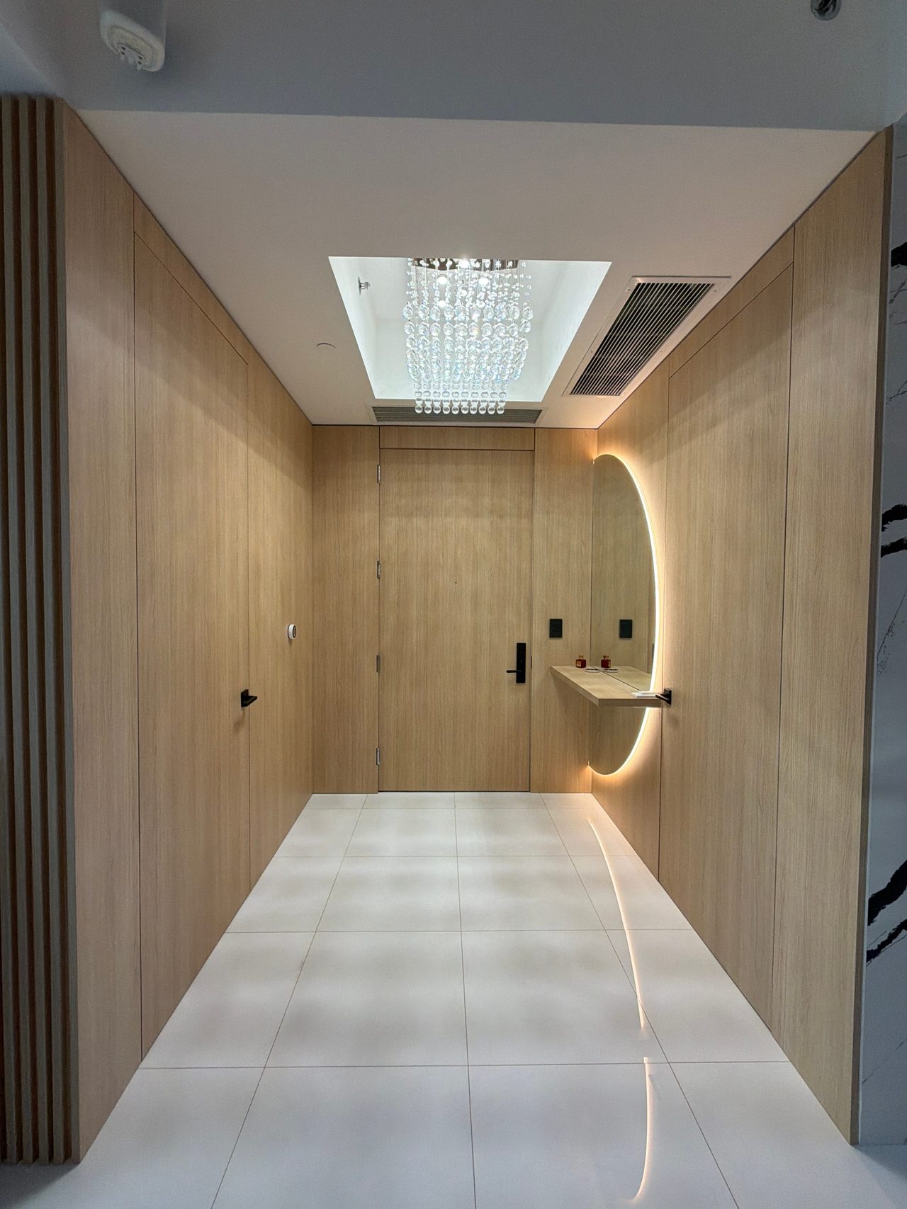 Narrow paneled entry hallway of light wood-grain flush doors leading to a backlit round mirror and a crystal ceiling fixture