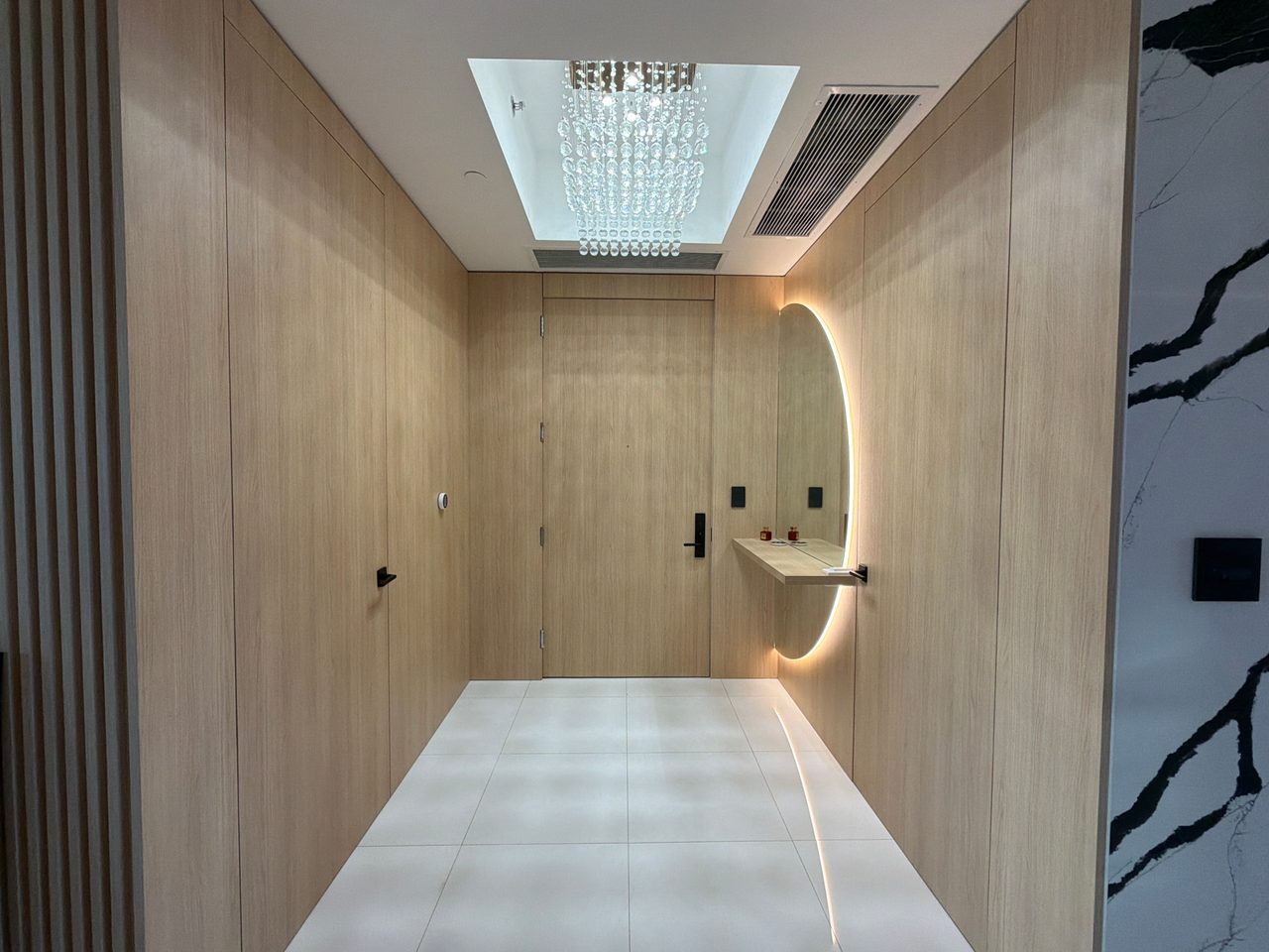 Paneled entry vestibule with flush wood-grain doors, a black lever handle, a backlit mirror, and a ceiling air grille beside a slatted wall