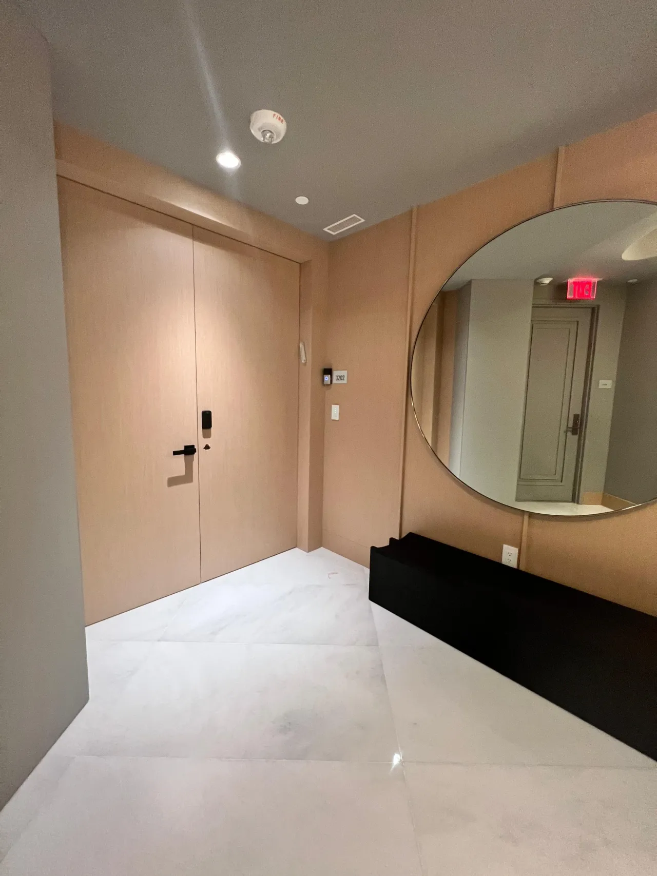 Pale wood-grain flush double entry door with a black lever handle, matching wall paneling, round wall mirror and light stone tile floor