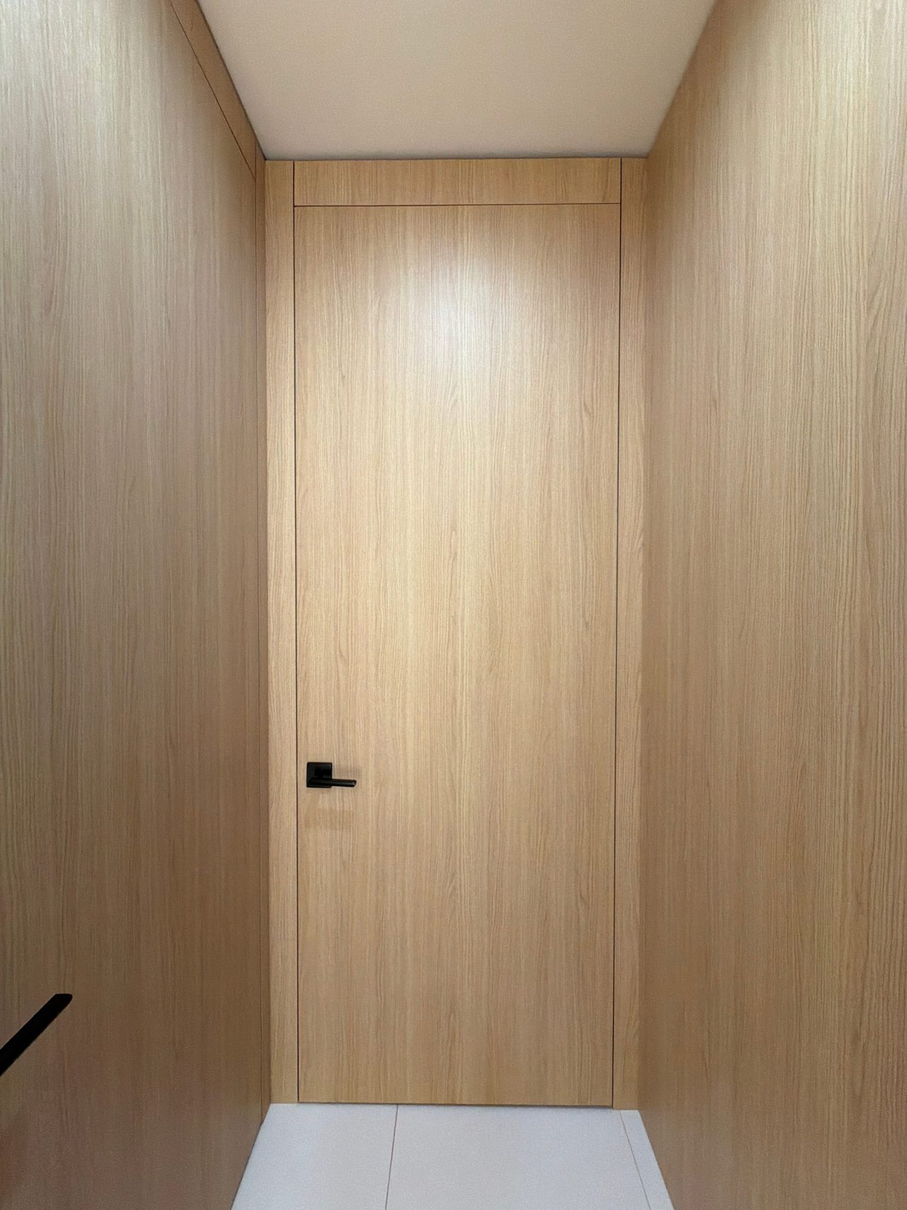 Tall light wood-grain flush door with a black lever handle set between matching full-height wall panels, with white tile flooring