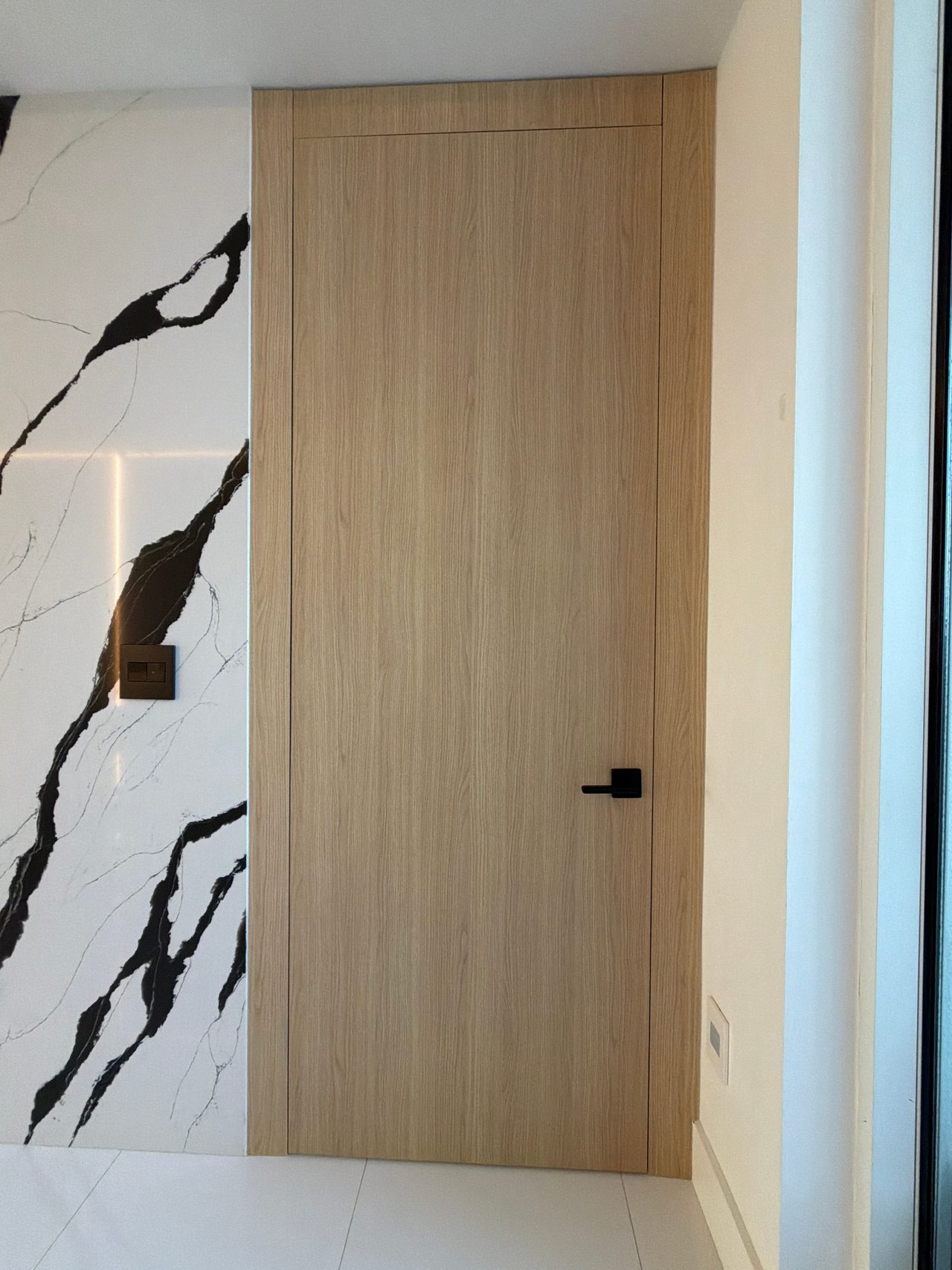 Tall light wood-grain flush door with a black lever handle beside a black-veined white stone-look wall and white tile floor
