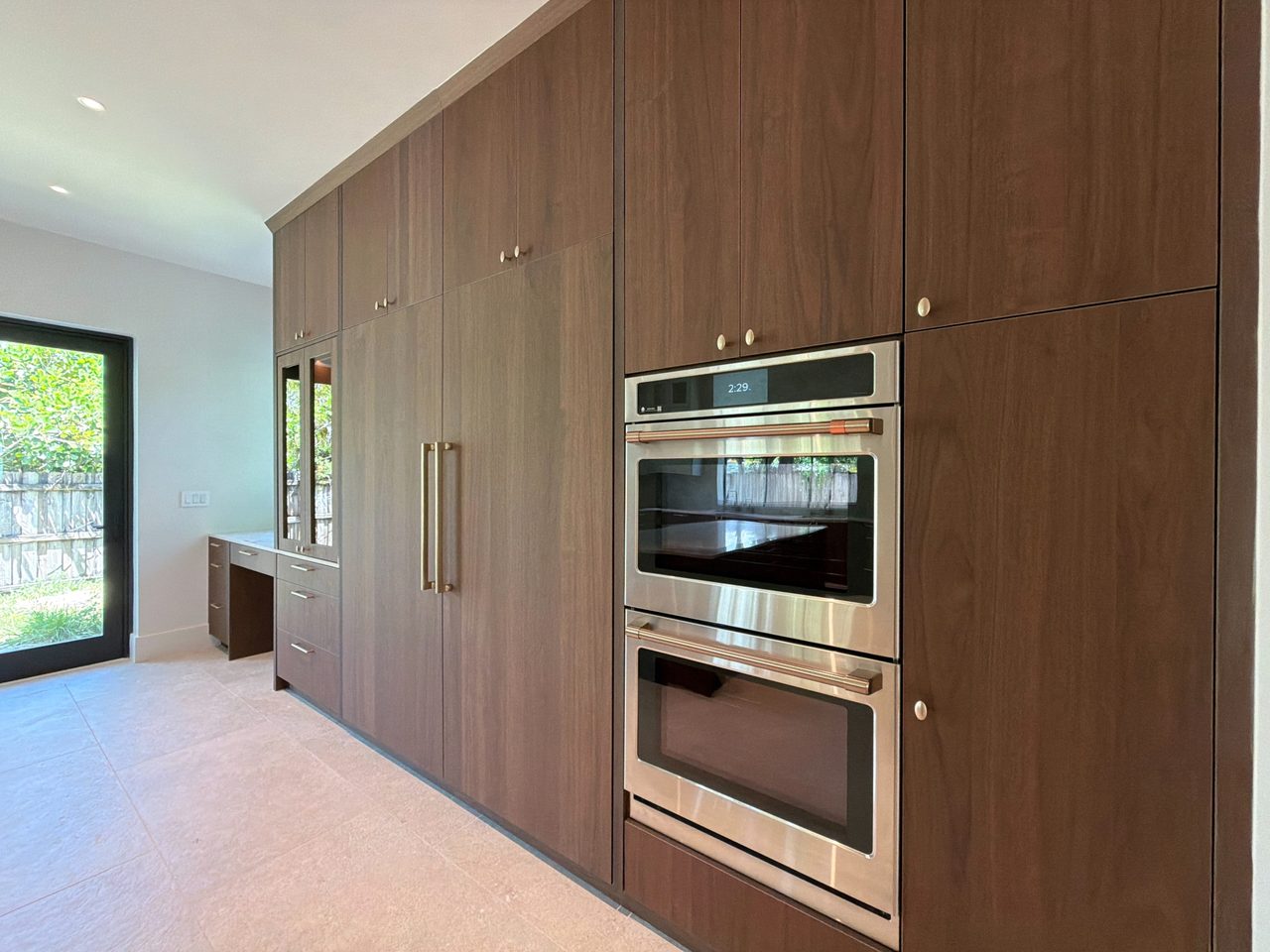 Angled view of a dark melamine cabinet wall with a stainless double wall oven and a glass-front cabinet, beside a desk nook and a glass patio door