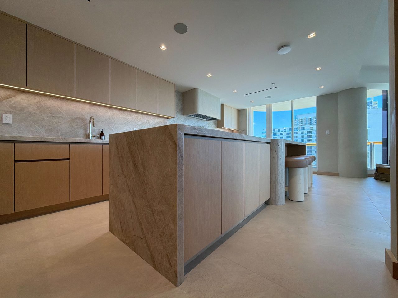 Angled view of a veined-stone waterfall island with a stone-clad column, leather-topped stools, and pale custom-stained wood veneer paneling