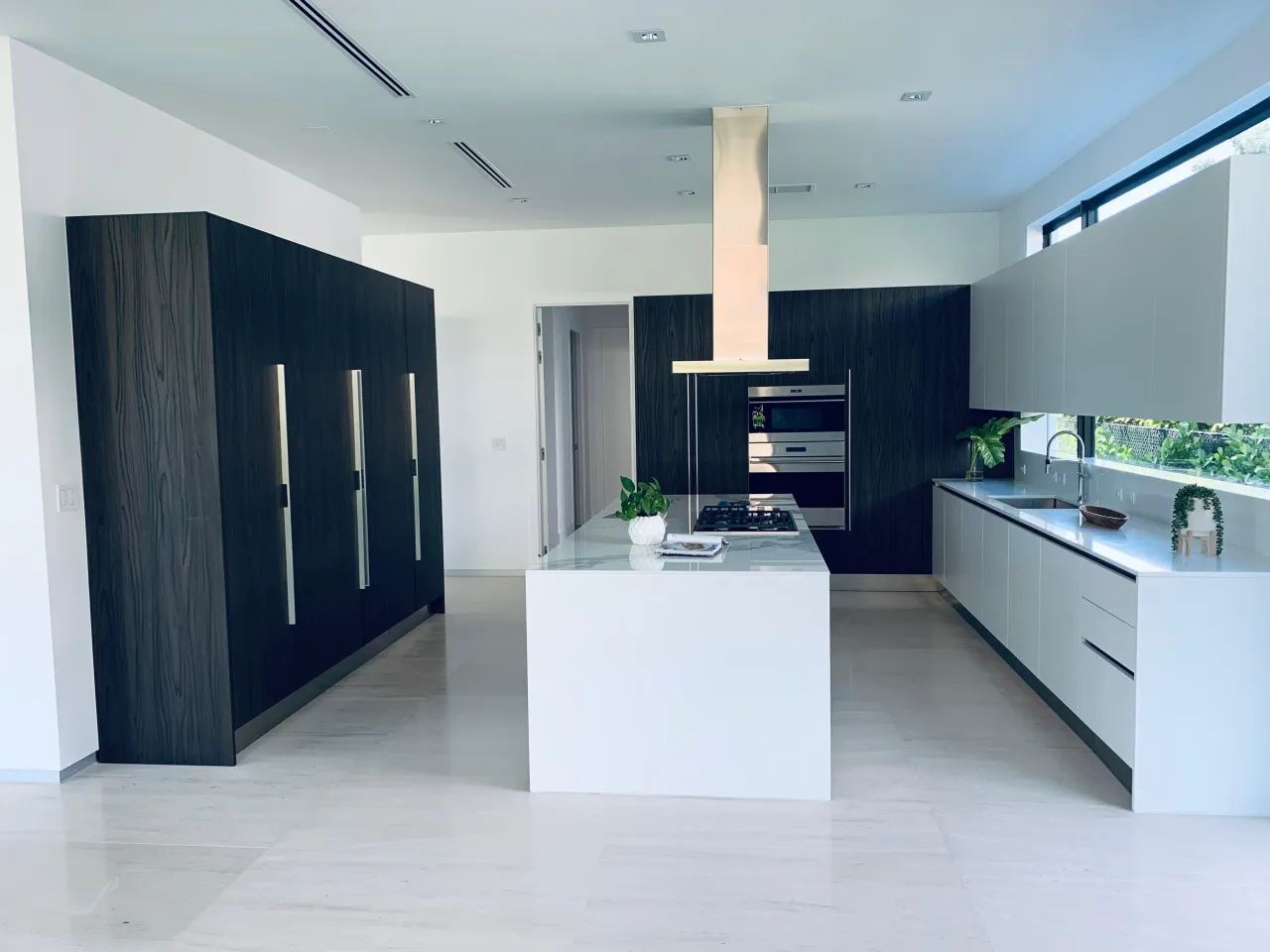 Black-and-white kitchen with a dark wood-grain tall cabinet block, light gray cabinets, and a white waterfall island with a gas cooktop and metallic hood
