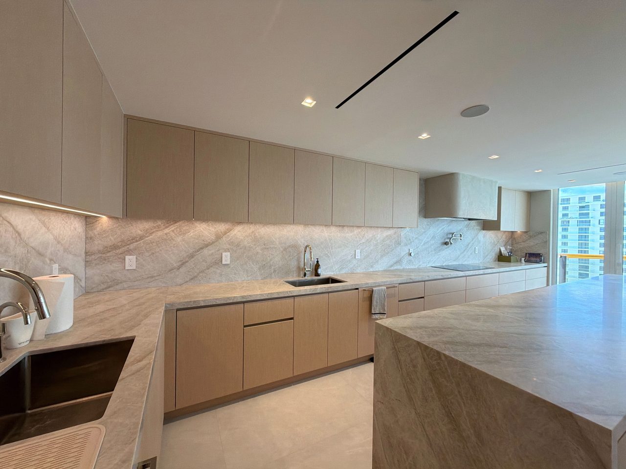 Custom-stained wood veneer lower cabinets under veined-stone counters wrapping a corner, with two undermount sinks and under-cabinet lighting