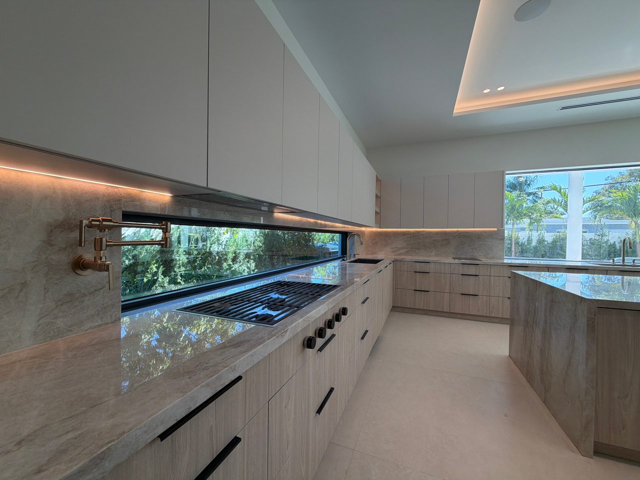 Kitchen counter with a gold-tone pot filler, veined-stone slab backsplash, a ribbon window over the cooktop, and light wood-grain melamine cabinetry