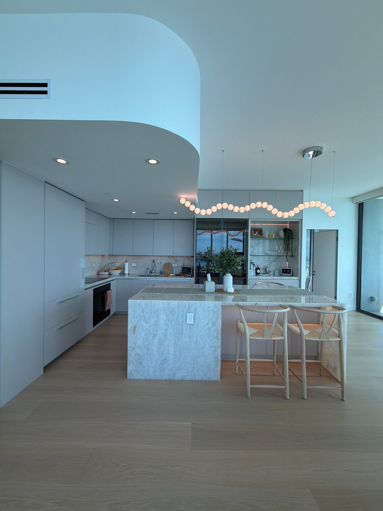 Kitchen with a curved white soffit, a strand of round pendant globes, a veined-stone waterfall island, and two woven-seat barstools