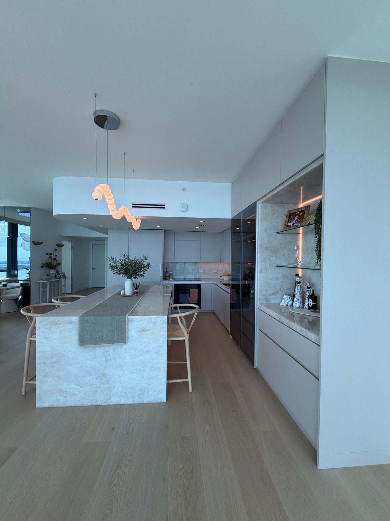 Kitchen with a curved white soffit, a veined-stone waterfall island, woven-seat stools, and a lit display niche with glass shelves on a stone back