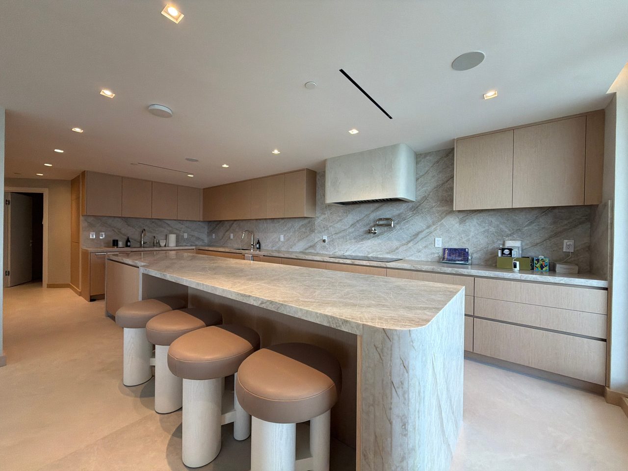 Open kitchen with custom-stained wood veneer cabinetry, a long veined-stone waterfall island with leather-topped stools, and a matching slab backsplash