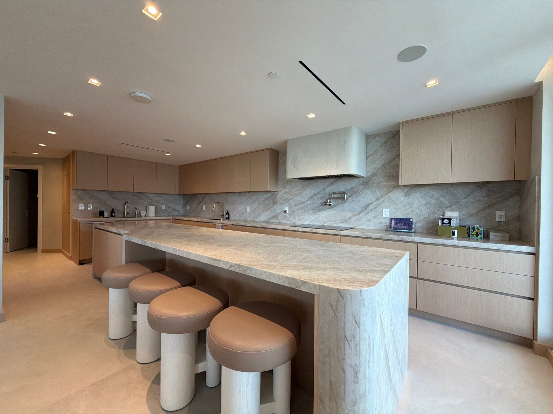 Open kitchen with custom-stained wood veneer cabinetry, a long veined-stone waterfall island with leather-topped stools, and a matching slab backsplash