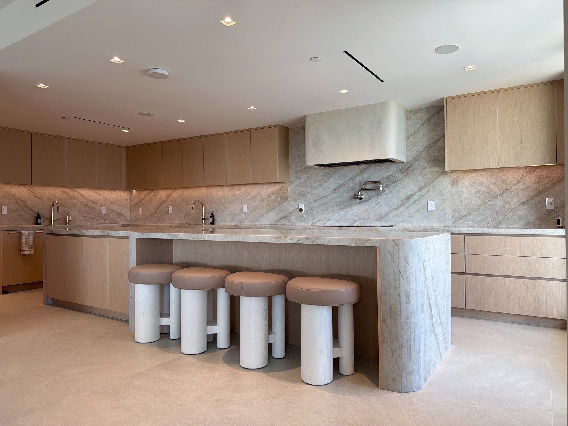 Open kitchen with pale custom-stained wood veneer cabinetry, a veined-stone waterfall island with leather-topped stools, and a slab backsplash and hood