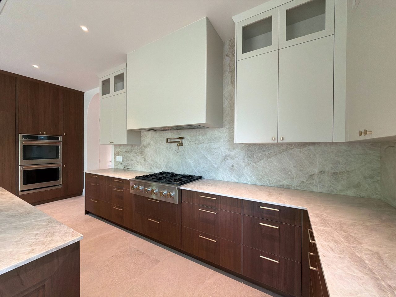Dark wood-grain melamine base cabinets with a gas cooktop and brushed-gold pot filler, cream uppers and hood, and a full-height veined-stone backsplash