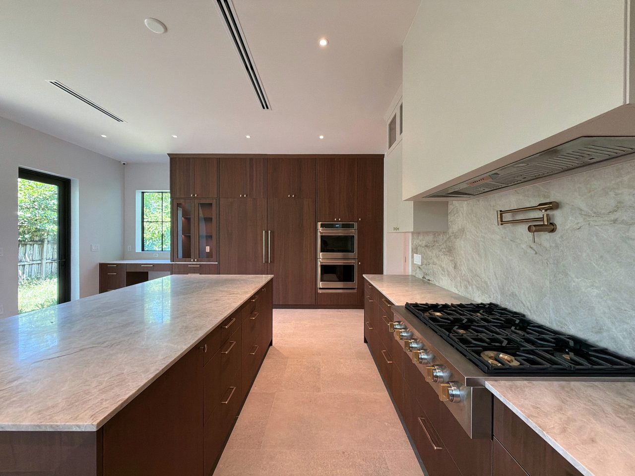Dark wood-grain melamine kitchen with a stainless gas rangetop, a brushed-gold pot filler on veined stone, a long island, and a tall oven cabinet