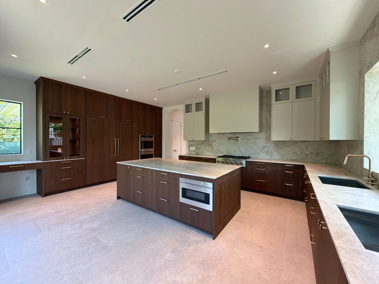 Dark wood-grain melamine kitchen with a glass-front display cabinet, a built-in desk nook, a veined-stone island, and cream upper cabinets
