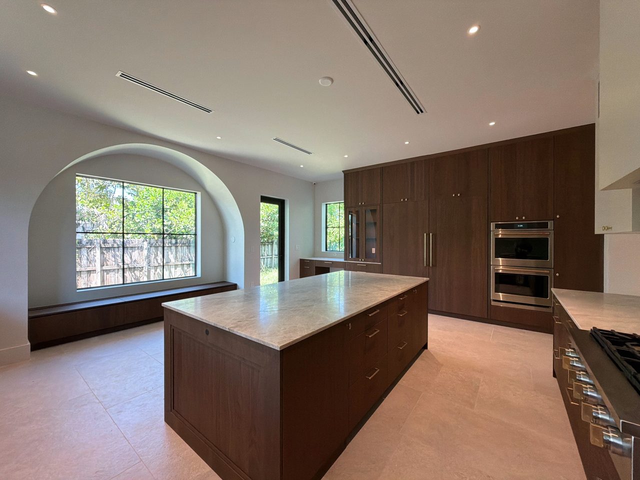 Dark wood-grain melamine kitchen from the breakfast area, with a veined-stone island, an arched steel-framed window over a bench, and a double wall oven