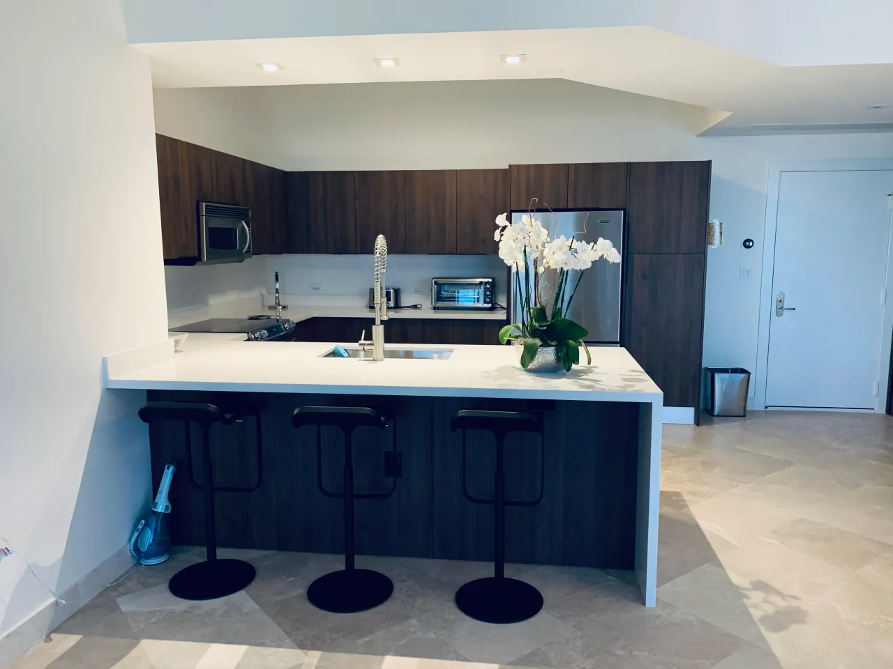 Kitchen with dark wood-grain cabinets, a white peninsula counter with three black stools, stainless microwave and refrigerator, and a stone tile floor