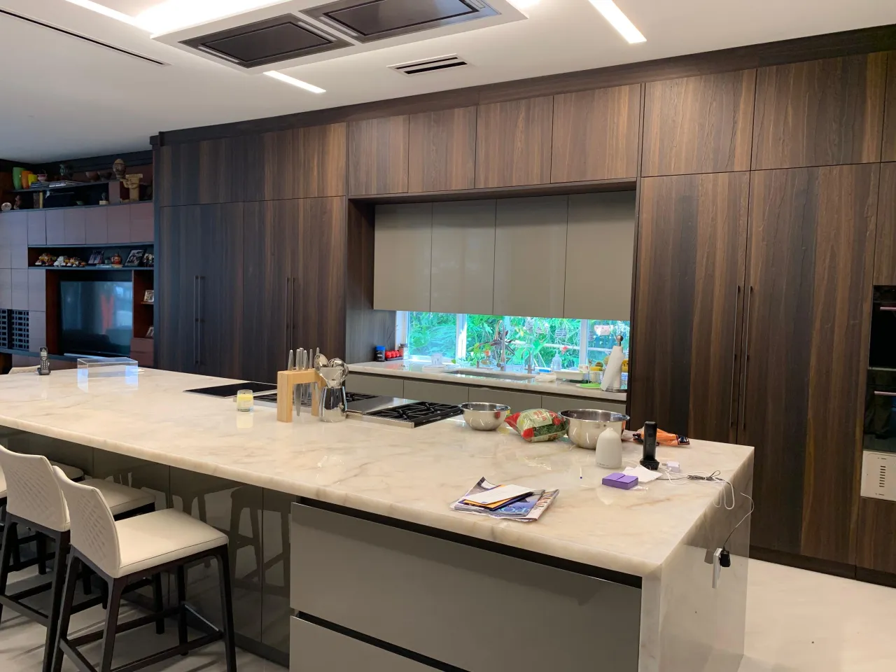 Dark wood-grain kitchen with a large veined-stone island, a black cooktop, recessed ceiling extractor panels, and white bar stools