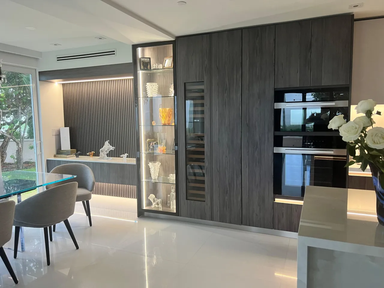 Dark wood-grain tall cabinet wall with a lit glass display case of glassware, a glass-front wine column, and stacked built-in wall ovens