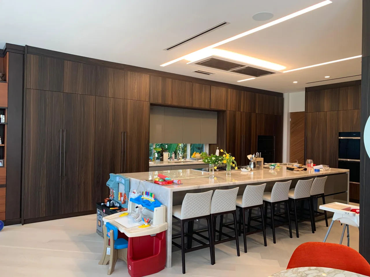 Dark wood kitchen with a large stone-top waterfall island, bar seating on quilted stools, linear LED ceiling coves, and built-in wall ovens