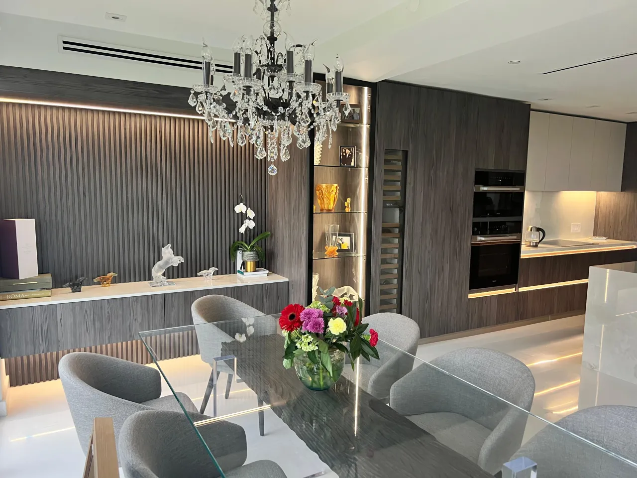 Dining/kitchen area with a crystal chandelier, a fluted wood wall panel, a glass wine storage column, and a glass dining table with upholstered chairs