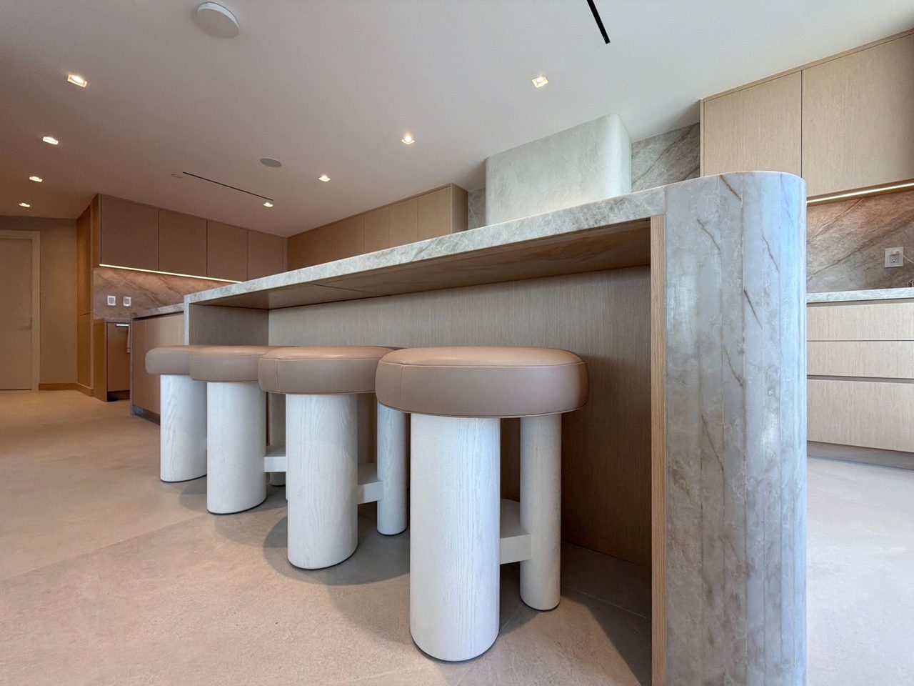 Four round leather-topped stools with pale wood pedestal bases tucked under the canted end of a veined-stone waterfall island