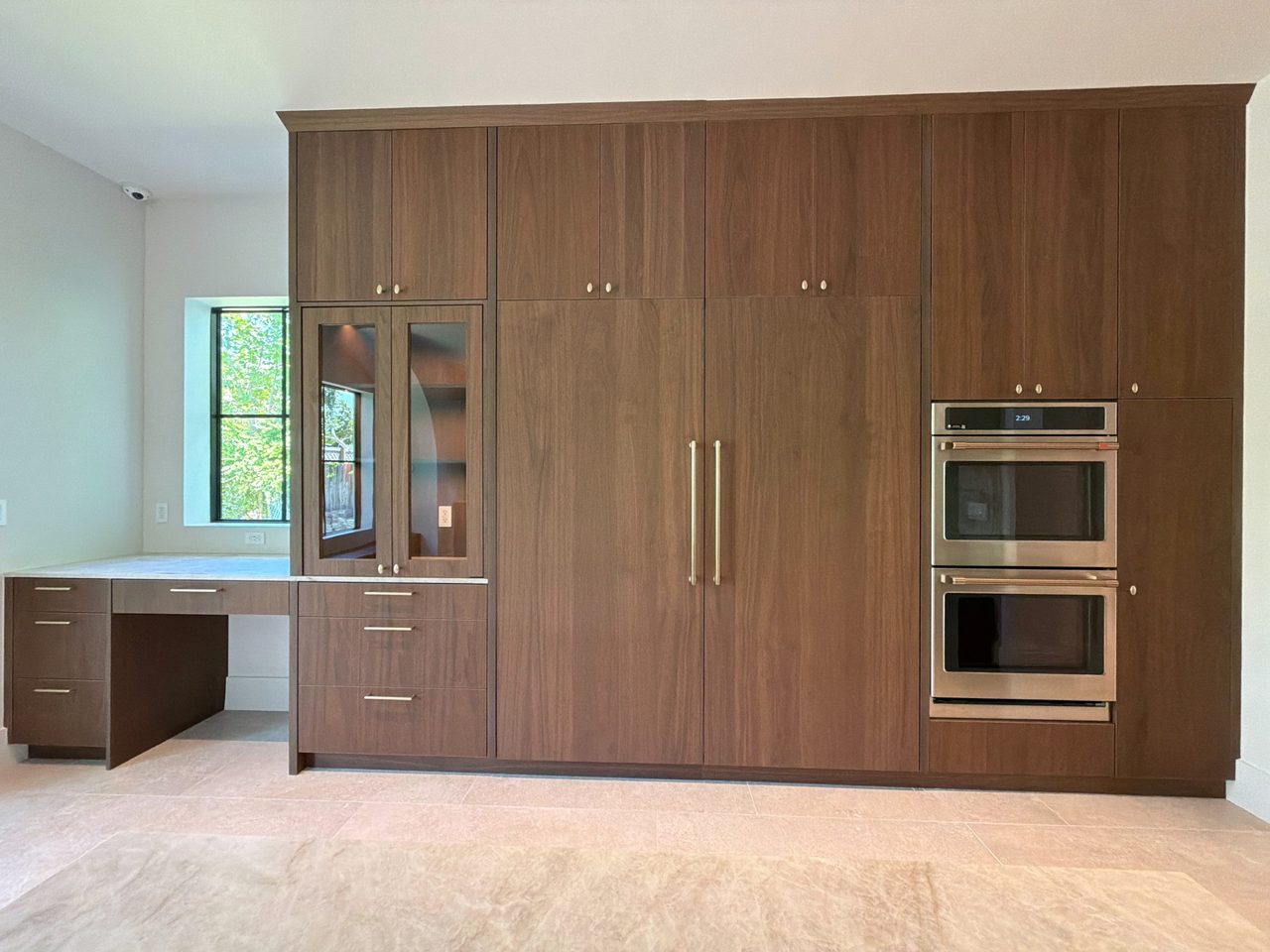 Full-height dark wood-grain melamine storage wall with a panel-front refrigerator, a stainless double wall oven, a glass-front display cabinet, and a desk