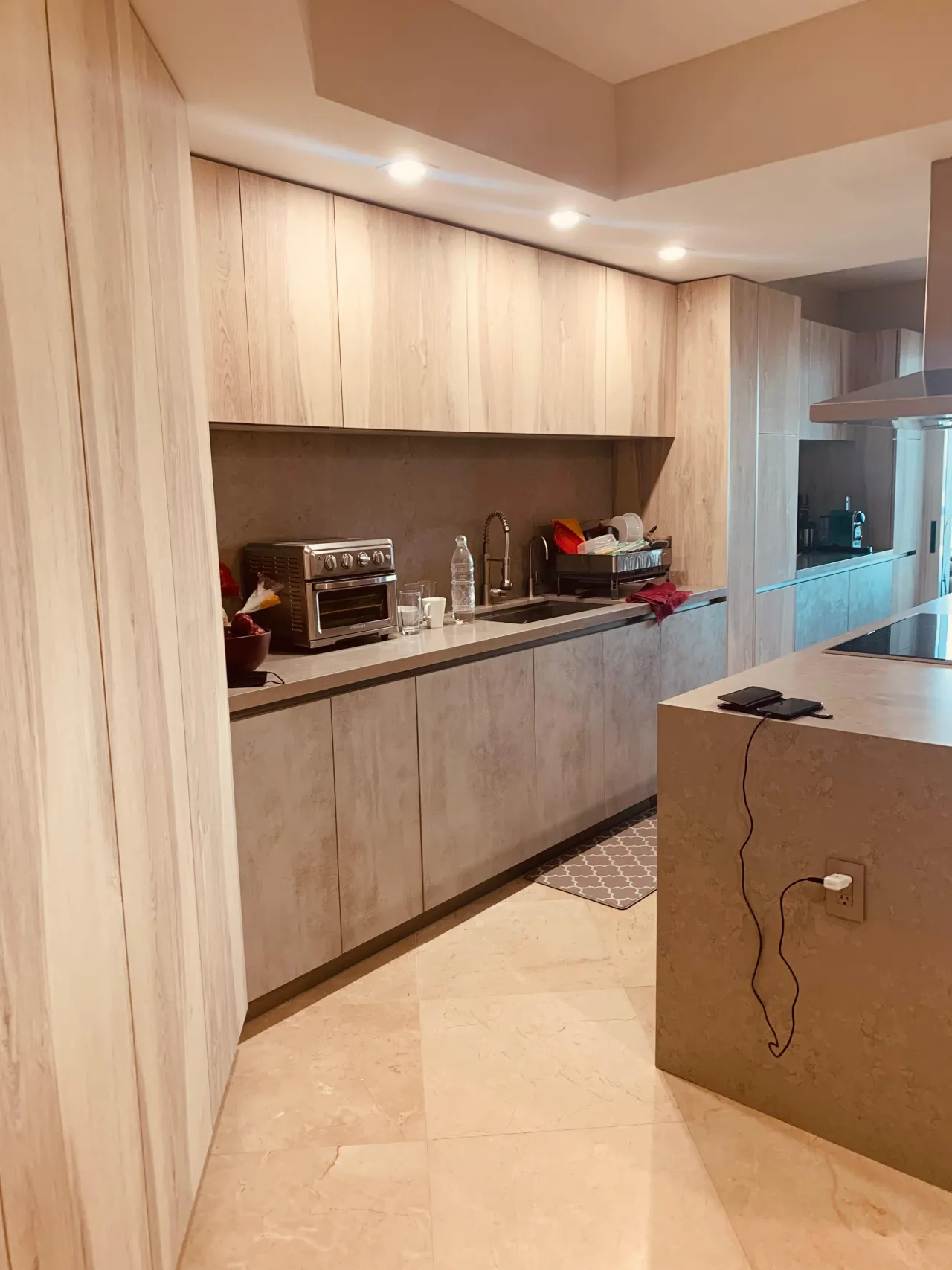 Galley kitchen with light wood-grain tall cabinets and uppers, gray concrete-look base cabinets and backsplash, stainless sink and stone tile floor
