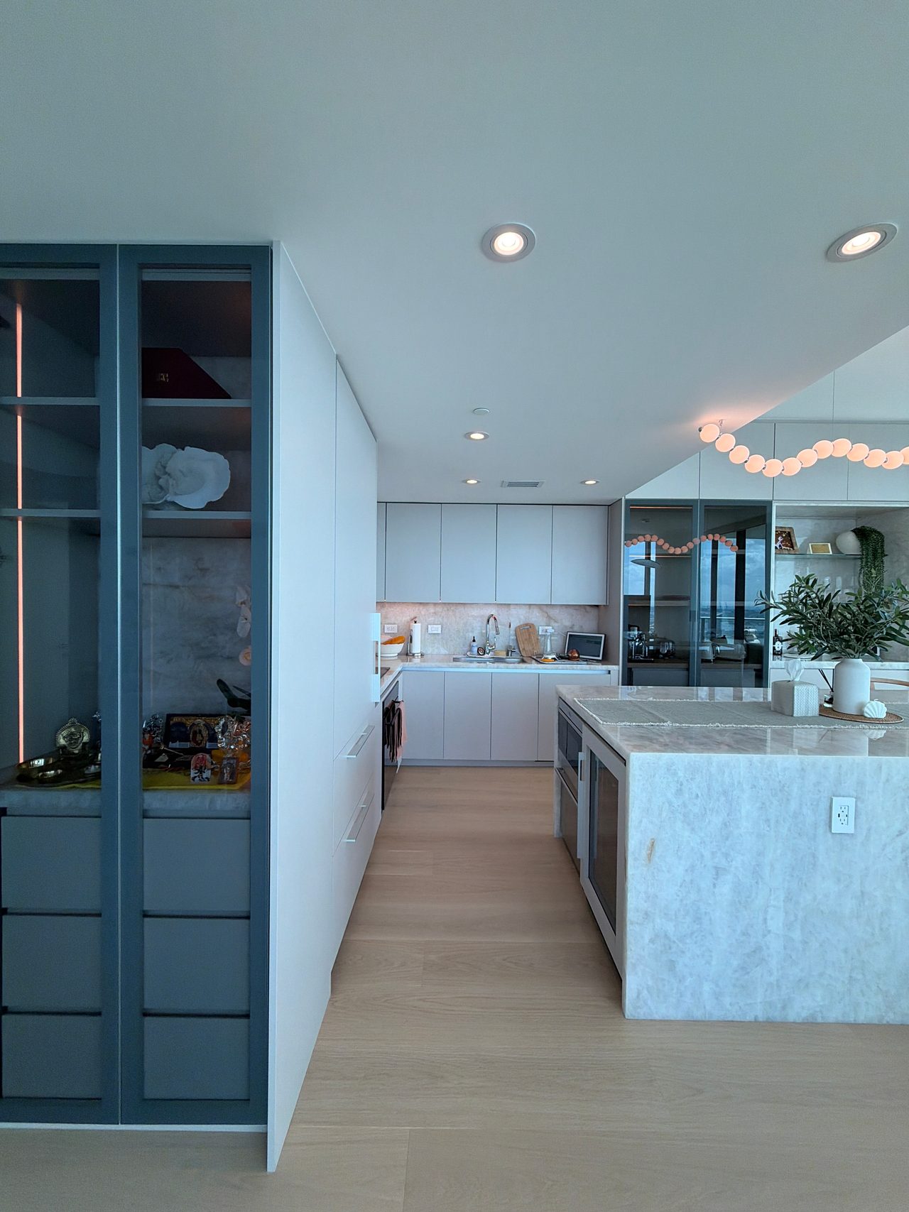 View down a kitchen past a glass-front display cabinet to a white cabinet run with a stone backsplash, and a veined-stone waterfall island