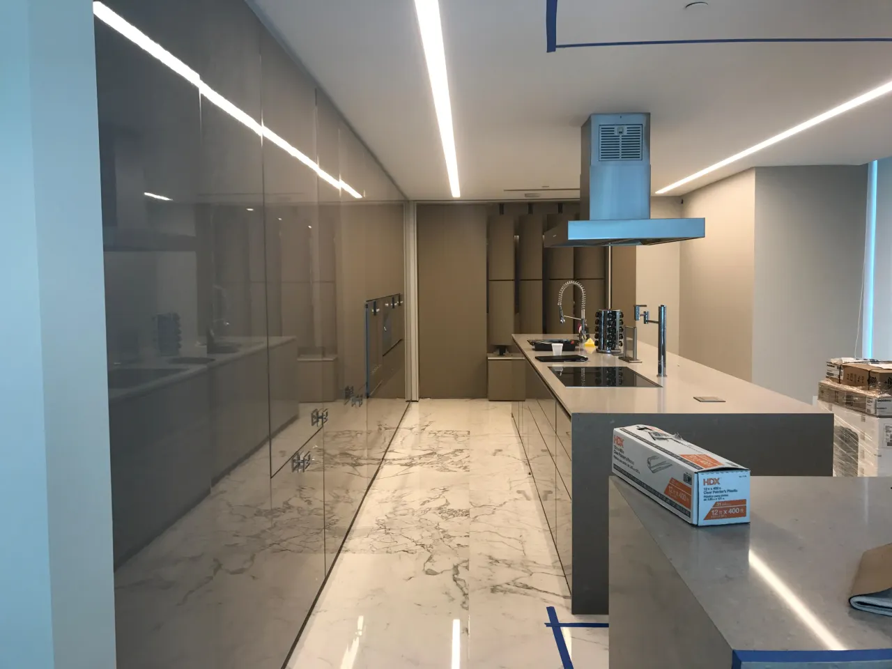 Glossy taupe/gray galley kitchen under construction with reflective glass-front cabinets, a white/gray stone tile floor, and a stainless hood