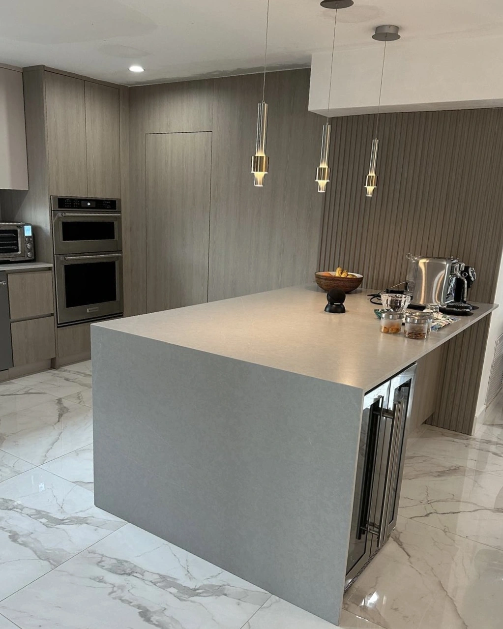 Gray stone waterfall island with a built-in beverage cooler, three slim pendants, wood-grain cabinets with double wall ovens and a veined stone floor