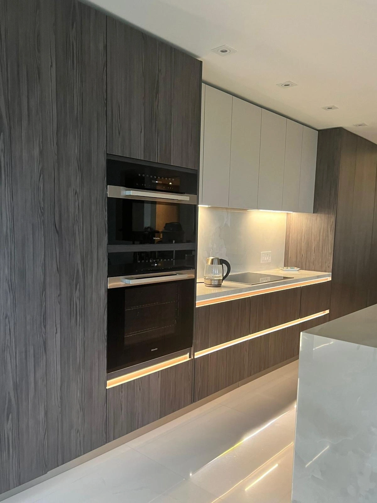 Gray wood-grain kitchen cabinets with a built-in double wall oven, LED under-cabinet lighting, and a light stone counter