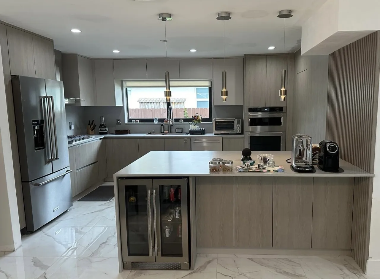 Gray wood-grain kitchen with a stainless French-door refrigerator, double wall ovens, a gas cooktop and a glass-front beverage cooler in the peninsula