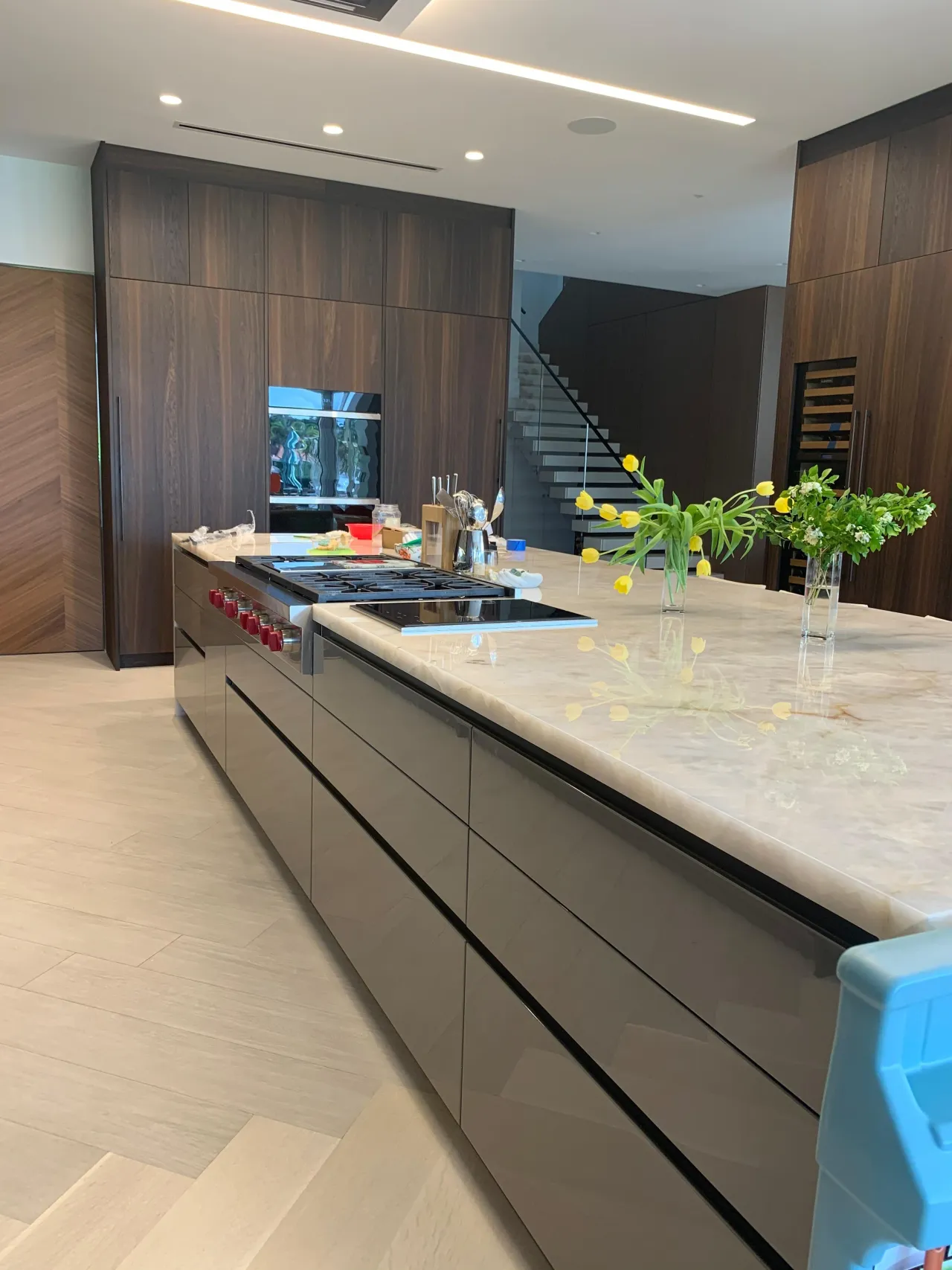 Island with glossy taupe fronts and a veined-stone top holding flowers, gas and induction cooktops, dark wood-grain wall cabinets, staircase beyond