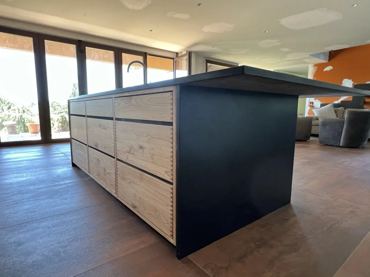 Kitchen island with pale dovetailed wood drawer fronts, a dark countertop and side panel, glass sliding doors behind and a gray polished floor
