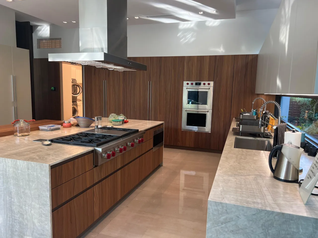 Kitchen island with a stainless gas cooktop with red knobs, a veined stone counter, a stainless island hood, and dark wood-grain cabinetry behind