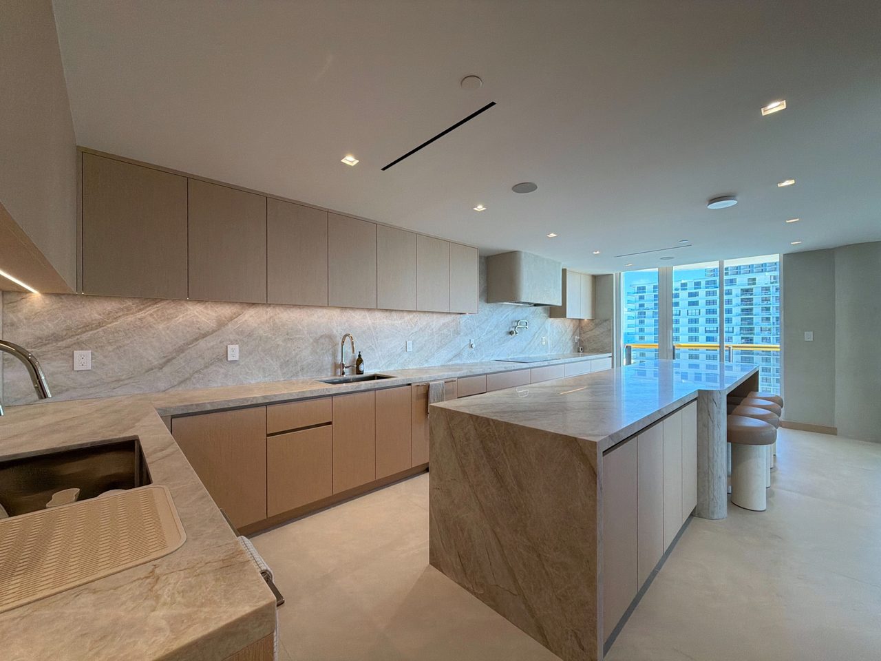 L-shaped pale wood veneer kitchen with two undermount sinks on a veined-stone counter and a waterfall island with round leather-topped stools