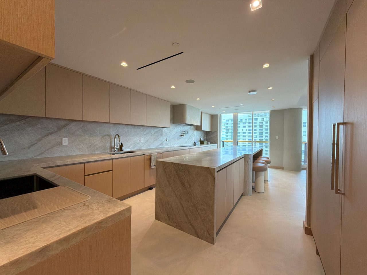 L-shaped kitchen with custom-stained wood veneer cabinets, veined-stone counters and backsplash, and a waterfall island by floor-to-ceiling windows