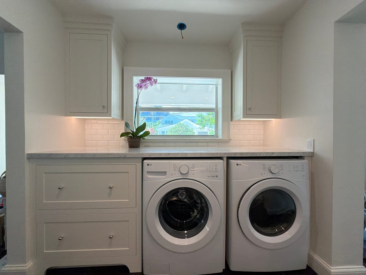 Laundry nook with white shaker drawer bases under a stone-look counter, front-load washer and dryer, flanking upper cabinets, and a window above
