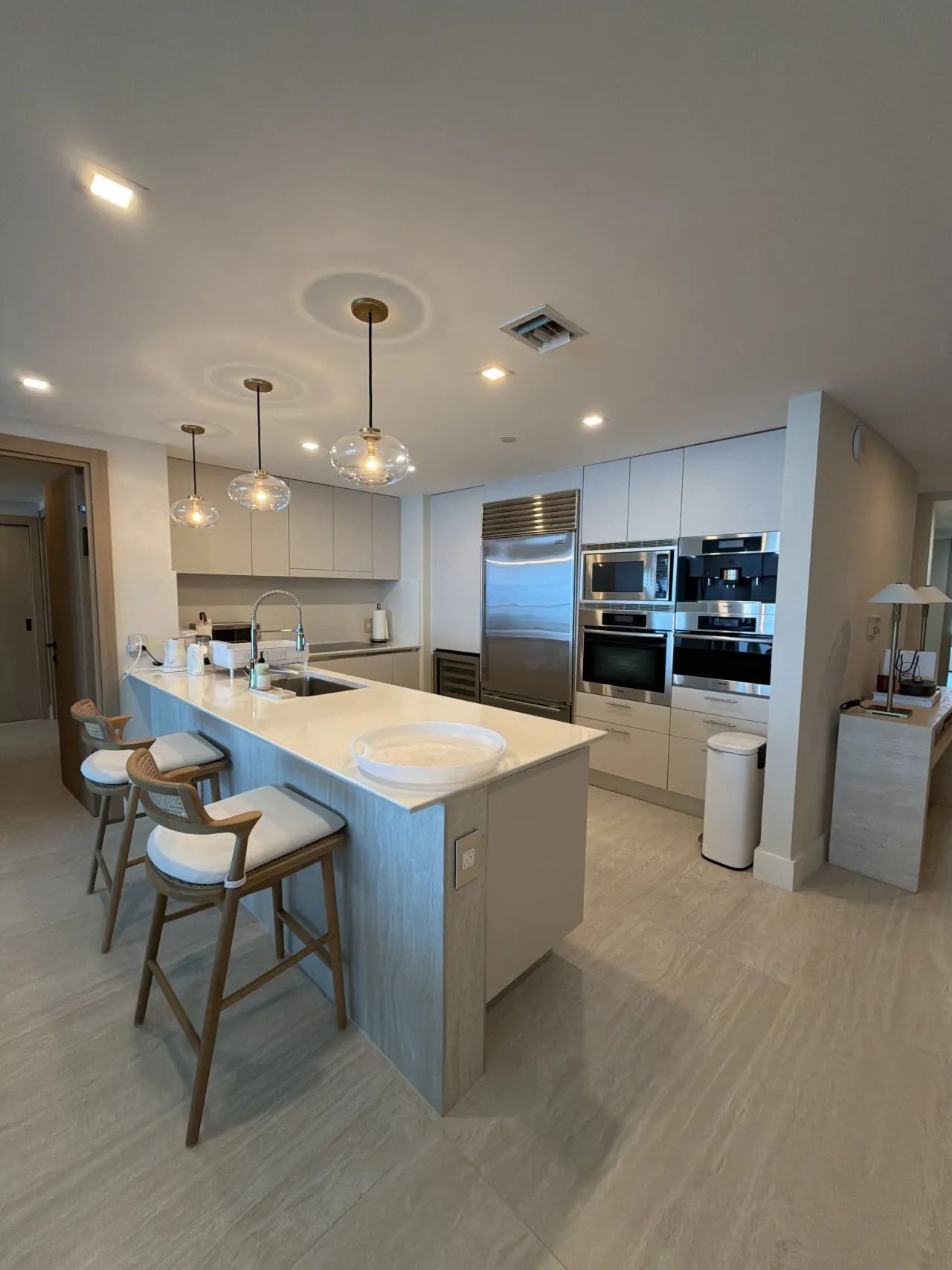Kitchen with a light stone-look peninsula and two barstools, three glass globe pendants, a stainless fridge, and stacked wall ovens
