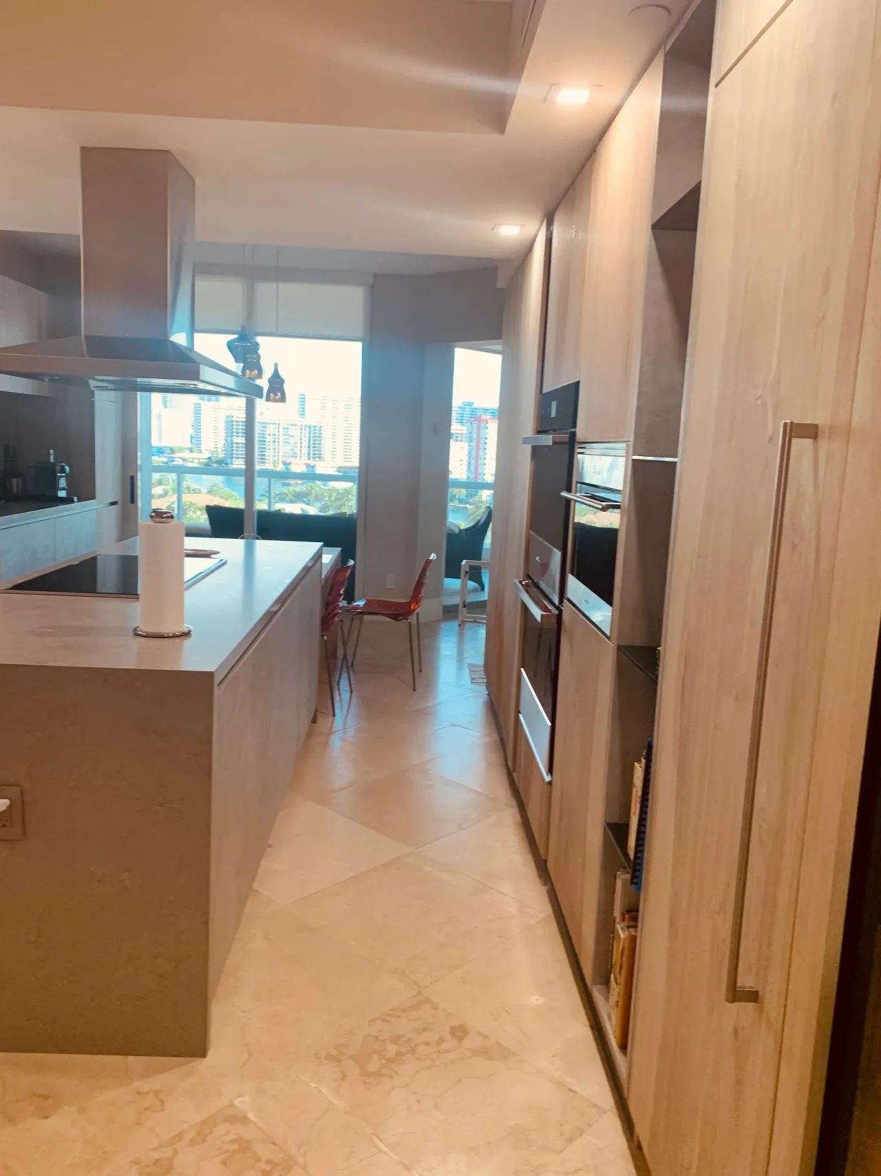 Light wood galley kitchen with built-in wall ovens, floor-to-ceiling windows showing a city skyline view, and a dining nook with red chairs