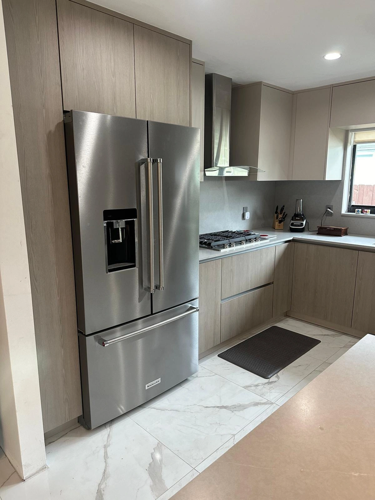 Kitchen with light wood-grain cabinets, a stainless French-door refrigerator, a gas cooktop under a stainless wall hood and a veined stone-look floor
