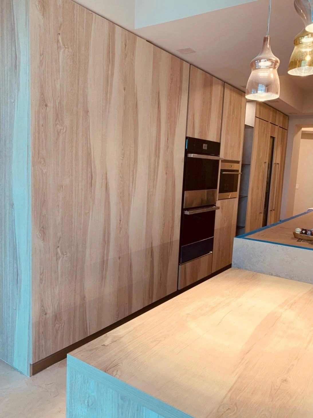 Light wood-grain floor-to-ceiling cabinet wall with two stacked built-in ovens, glass pendant lights, and a wood-topped island in the foreground
