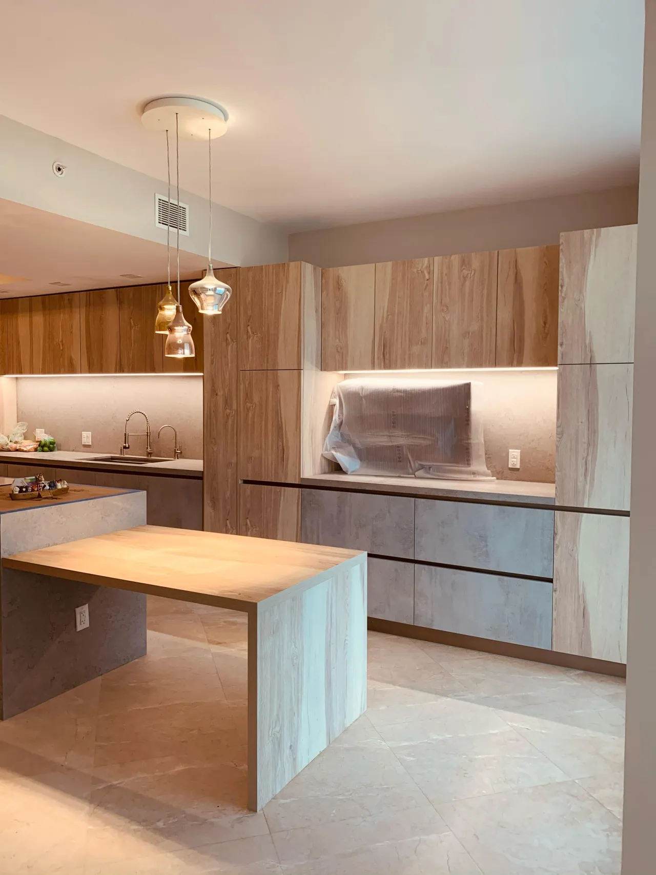 Light wood-grain kitchen with concrete-look drawer fronts, a plastic-wrapped hood in a lit niche, three glass pendants, and a wood table off the island