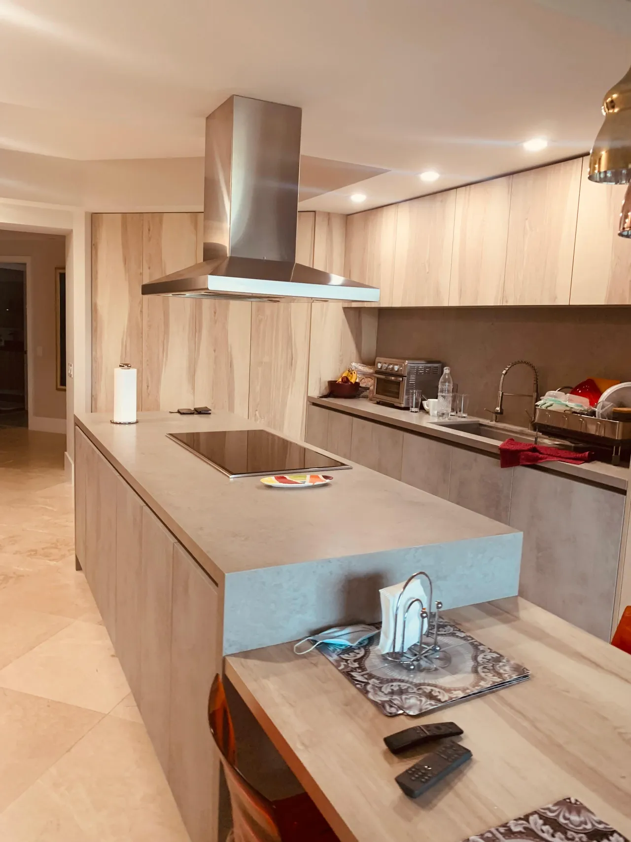 Light wood-grain kitchen island with a stainless range hood over a black cooktop, adjoining a wood dining table
