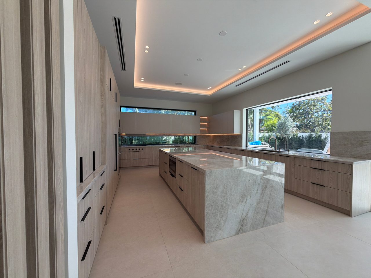 Kitchen in light wood-grain melamine with a veined-stone waterfall island, black bar pulls, and a wide picture window over the counter showing greenery