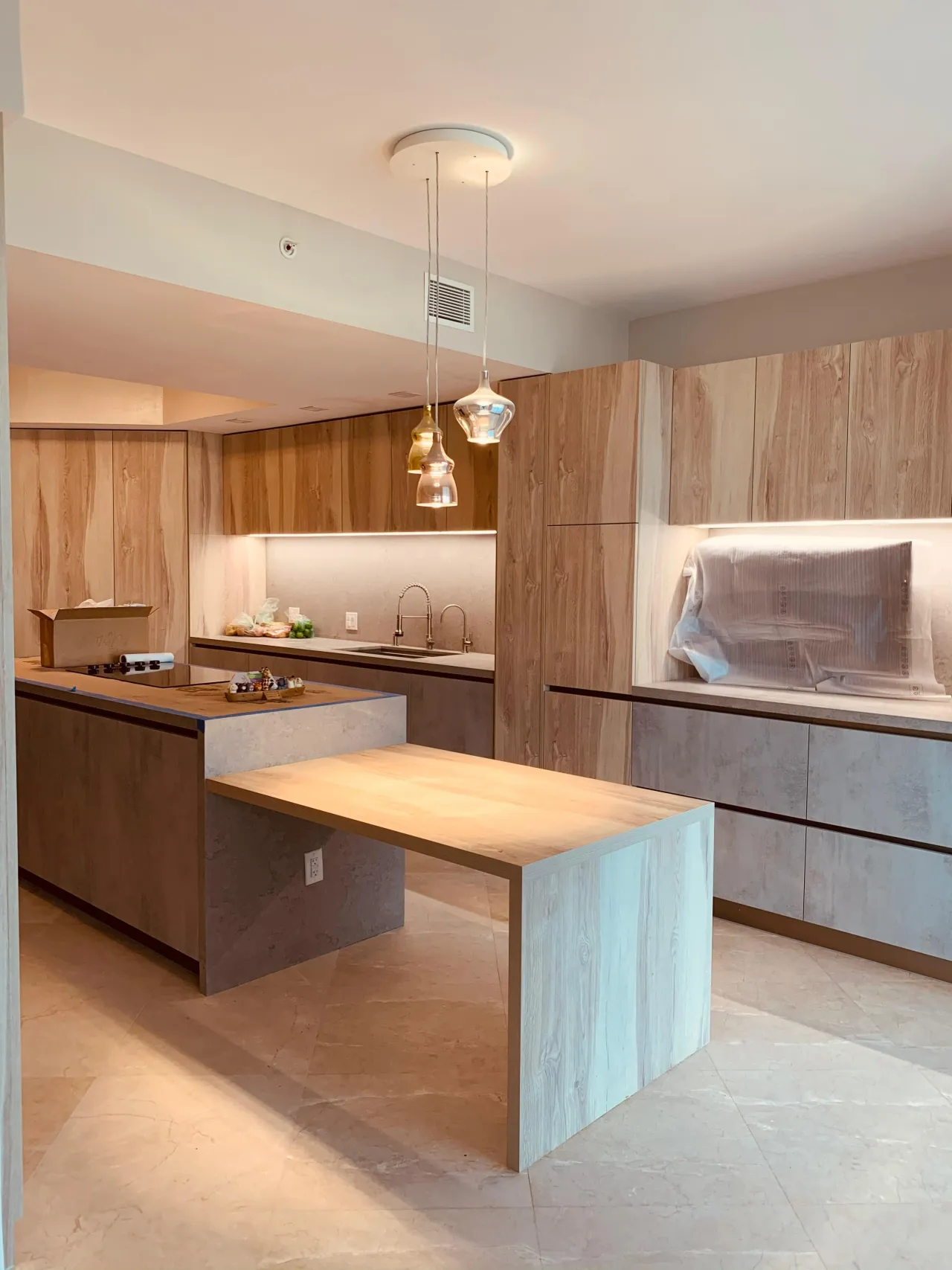 Light wood-grain tall cabinets and stone-look base units, with two glass pendants above a wood table extending from the stone-look island
