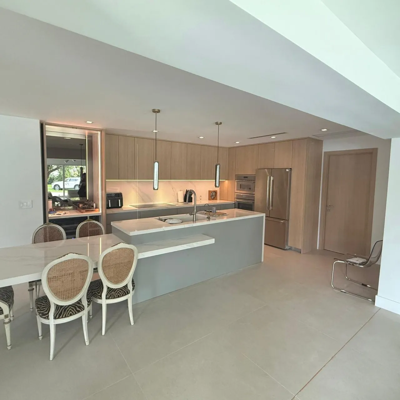 Light wood kitchen with a two-tier gray and veined-stone waterfall island, pendant lighting, and a stainless panel refrigerator with a built-in wall oven