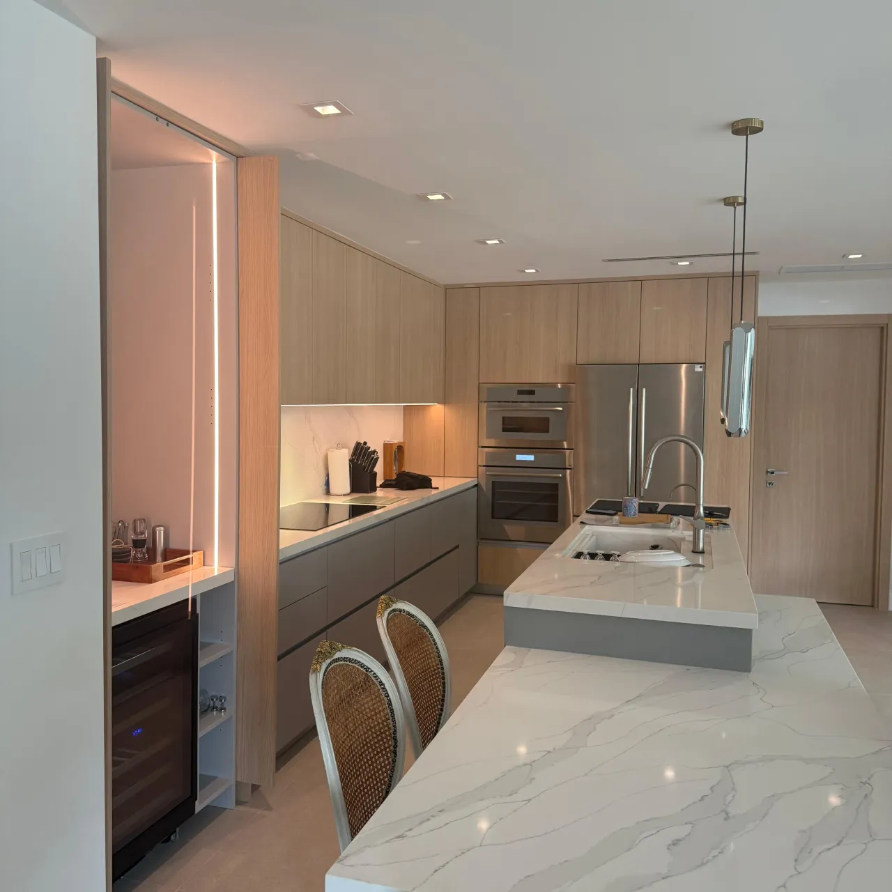 Light wood kitchen with a veined-stone waterfall island featuring an integrated sink and cooktop, pendant lighting, and stainless double wall ovens