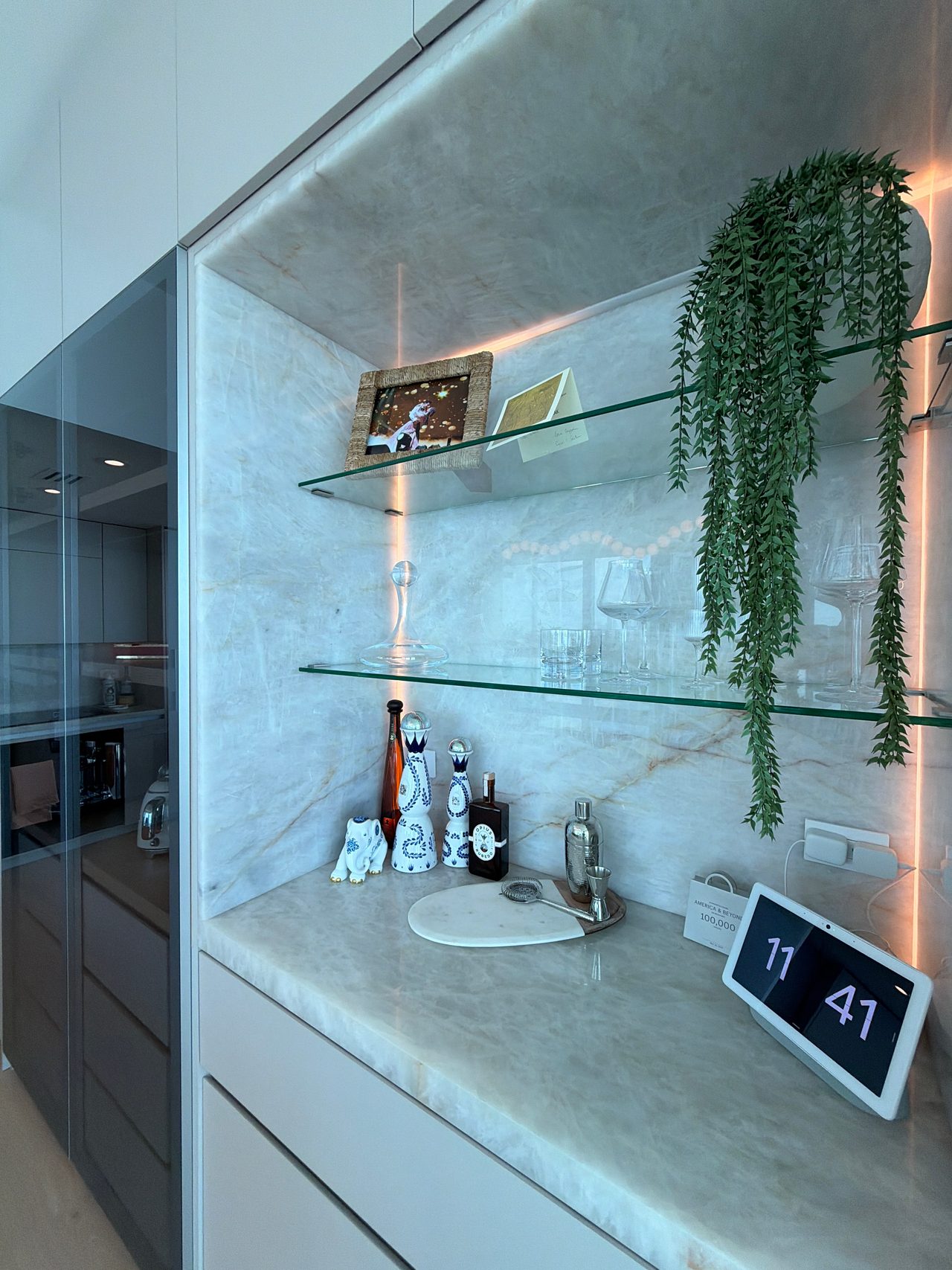 Lit display niche set into a veined-stone wall with two glass shelves of glassware and decor, a counter below, and a hanging plant